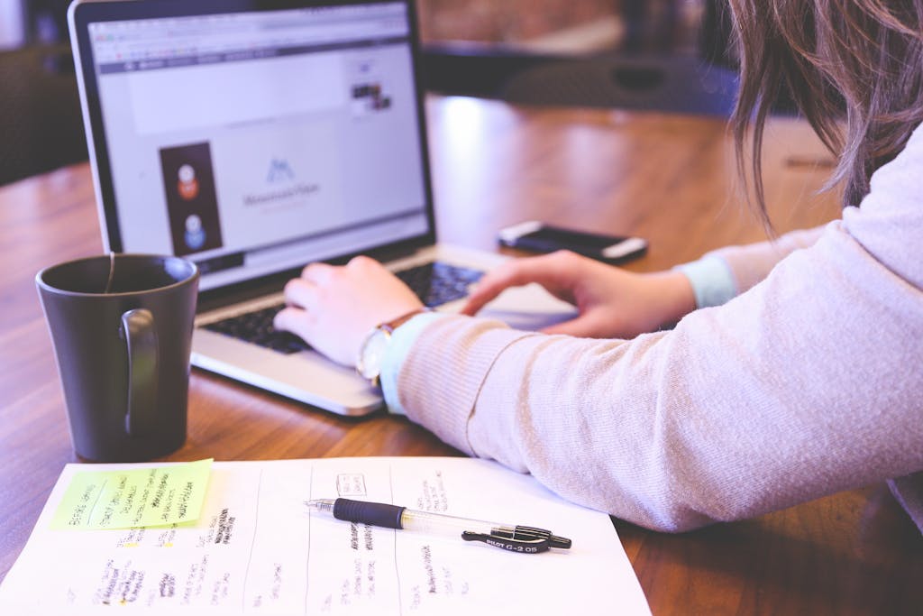 Businesswoman typing on laptop with notes and coffee on wooden desk.
